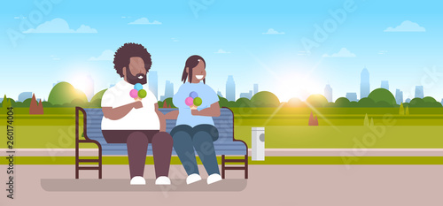 fat obese couple sitting wooden bench eating ice cream overweight african american man woman having fun relaxing in city park sunset cityscape background full length flat horizontal