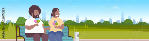 fat obese couple sitting wooden bench eating ice cream overweight african american man woman having fun relaxing in city park sunset cityscape background portrait flat horizontal