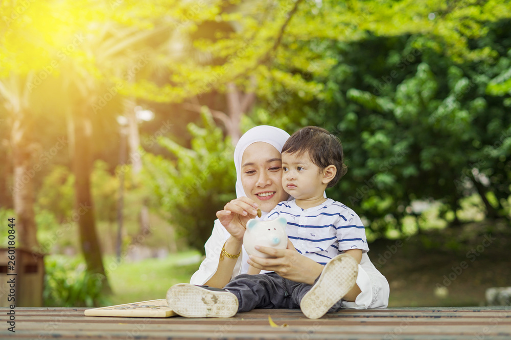 Muslim Asian young mother with her baby son, learning and putting coins into piggy bank at park. Education, financial and future planning concept.