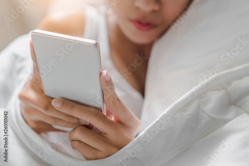 Close up of young asian woman playing with her smathphone on white pillow on bed in bedroom on morning time after wake up, Selective focus.