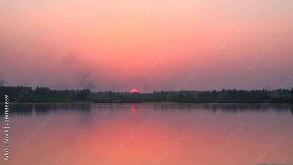Time lapse of sun setting in a brilliant colorful red and orange sky with reflection in the water across the lake and forest somewhere on the highway in Karnataka, India Time lapse of sun setting in a brilliant colorful red and orange sky with reflection in the water across the lake and forest somewhere on the highway in Karnataka, India