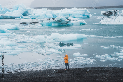 Young girl standing on the sea shore covered in ice floes taking picture with mobile phone of the Nordic landscape in Iceland