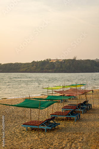 empty blue beach beds under color awnings on the yellow sand against the background of the sea and the green island with the house in the evening