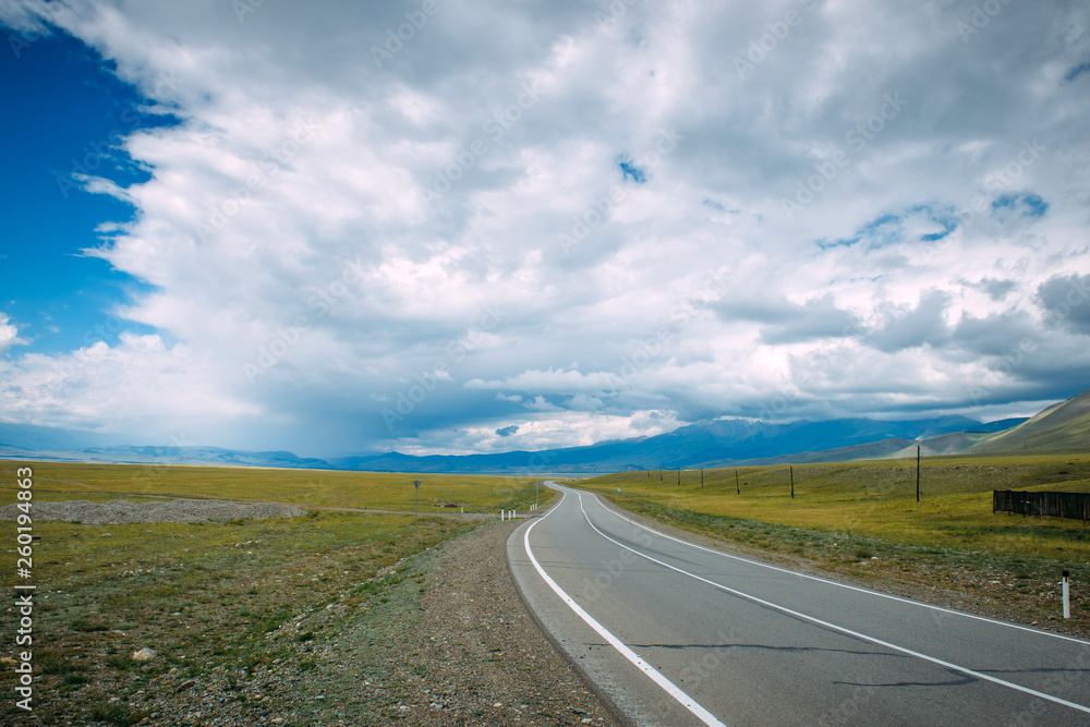 A winding road running in a mountainous area. Smooth asphalt road passes between the yellow plain to the distant mountains. The Chui highway through the kurai steppe, Altai. Low clouds over the road. A winding road running in a mountainous area. Smooth asphalt road passes between the yellow plain to the distant mountains. The Chui highway through the kurai steppe, Altai. Low clouds over the road.