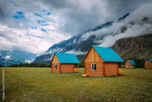 Wooden houses in a green valley on a background of mountains in the fog and cloudy sky, close-up. Tourist base in the Altai mountains. Houses near the big rock, summer morning landscape.