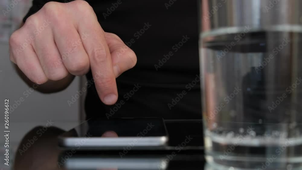 Man's finger controlling smartphone on a table with a glass of water on the foreground. Man's finger controlling smartphone on a table with a glass of water on the foreground.