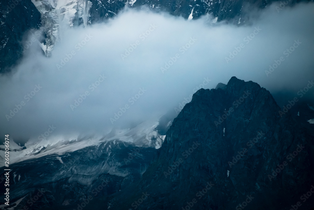 Low cloud before huge glacier. Giant snowy rocky mountain wall in thick fog. Early morning in mountains. Impenetrable fog. Dark atmospheric foggy landscape with cold rocks. Tranquil mystic atmosphere.
