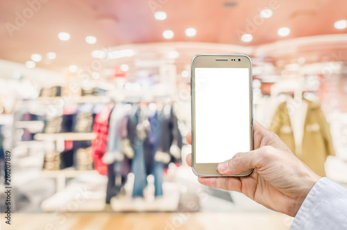 Mockup image of man hand holding gold mobile phone with blank white screen on blurred abstract background of many cotton clothing on the shelves of fashion shop.business concept.shopping online.