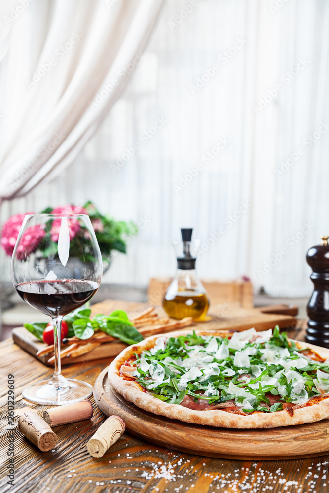 Close up view on fresh served pizza with prosciutto on a wooden table on light background. Pizza with wine. Vertical image with copy space for text. Food for menu. Italian cuisine. 