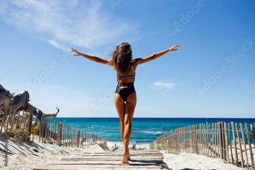 Rear view of a beautiful, brunette young girl with raised hands, looking at ocean. Freedom concept, holiday, beach, clear sky background., light and shadow landscape scenery place