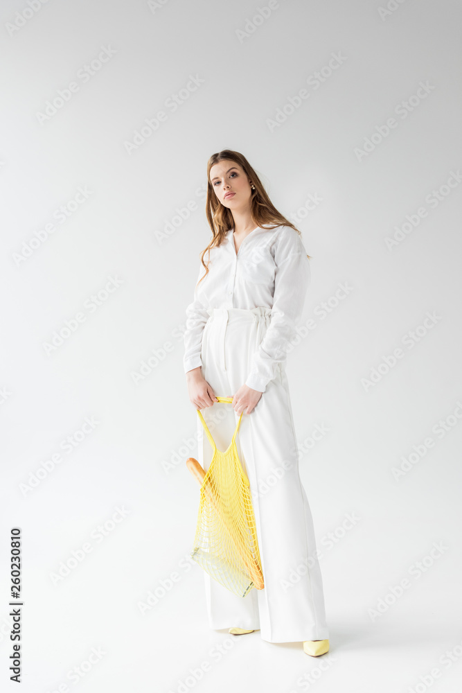 pretty young woman holding yellow string bag with baguette and bottle of milk on white