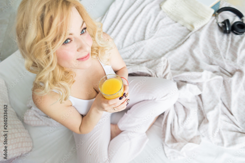 Attractive blonde woman sits on the edge of the bed at home and holds a stocker with orange juice in her hand. Morning mood and vigor Attractive blonde woman sits on the edge of the bed at home and holds a stocker with orange juice in her hand. Morning mood and vigor