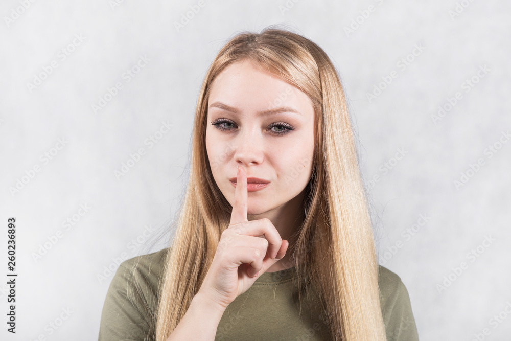 Portrait of a young beautiful woman showing a gesture of silence with a finger to his lips on a gray background. Body language concept. Portrait of a young beautiful woman showing a gesture of silence with a finger to his lips on a gray background. Body language concept.