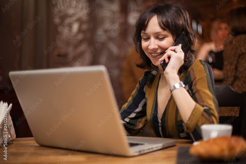 Beautiful businesswoman working on her laptop while having a conversation on the phone in a coffee shop Beautiful businesswoman working on her laptop while having a conversation on the phone in a coffee shop
