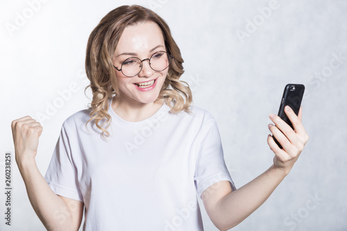Happy young woman in glasses holding smartphone and celebrating his success.