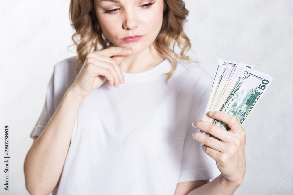 Pensive young woman looks at bunch of money, thinks where to spend it, over white background. Pensive young woman looks at bunch of money, thinks where to spend it, over white background.