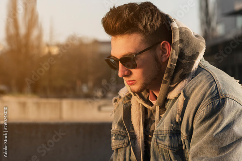 Fashionable handsome hipster man model with stylish sunglasses sitting and thinking on a bench in the city at sunrise.