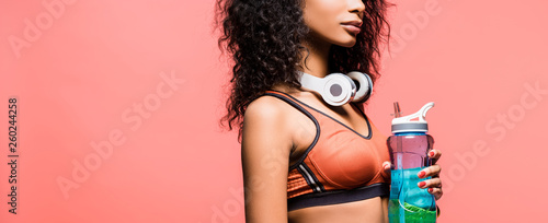 panoramic shot of african american sportswoman with headphones and sport bottle isolated on coral with copy space