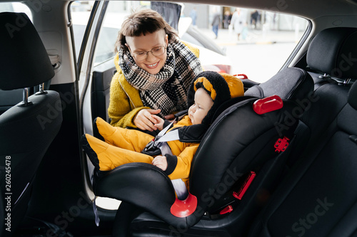A pretty brunette in a yellow jacket fastens the child car seat belt before going for a walk with her baby. © galaganov