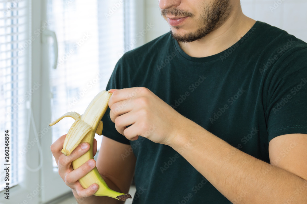 A young handsome brunette man is standing in the kitchen at home and cleans a banana. A young handsome brunette man is standing in the kitchen at home and cleans a banana.
