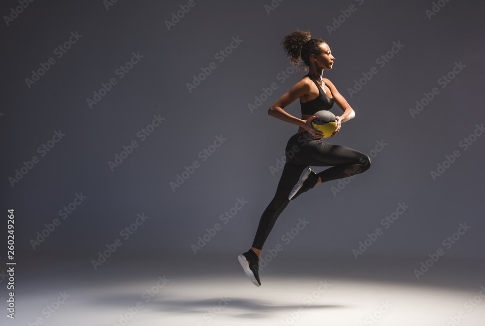 side view of athletic african american sportswoman holding ball and jumping on grey