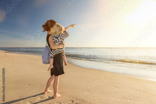 Mutter mit Kind auf dem Arm steht am Strand und schaut in den Sonnenuntergang