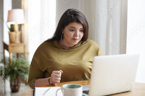 Attractive young dark haired woman with curvy body using generic laptop for remote work, drinking coffee, looking at screen with concentrated focused facial expression. Technology and lifestyle