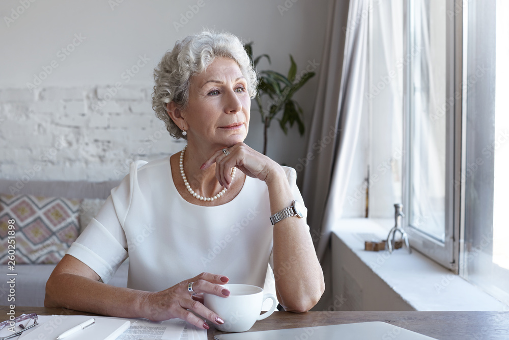 Attractive classy sixty year old mature woman wearing stylish wrist watch and pearl necklace spending nice time at home alone, sitting at her workplace with cup of coffee, looking through window Attractive classy sixty year old mature woman wearing stylish wrist watch and pearl necklace spending nice time at home alone, sitting at her workplace with cup of coffee, looking through window