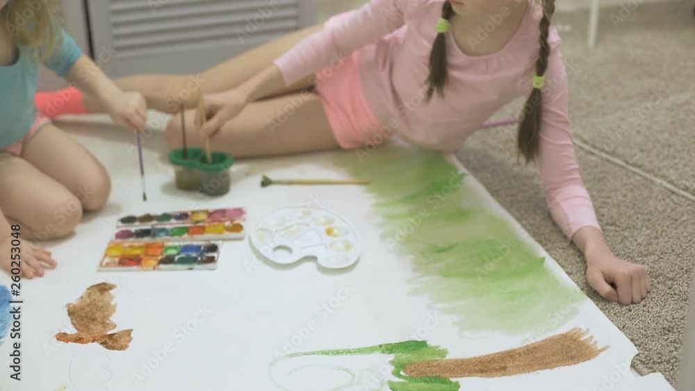 two girls paint with watercolor paints and brushes on a large drawing paper on the floor , children's joint creativity two girls paint with watercolor paints and brushes on a large drawing paper on the floor , children's joint creativity