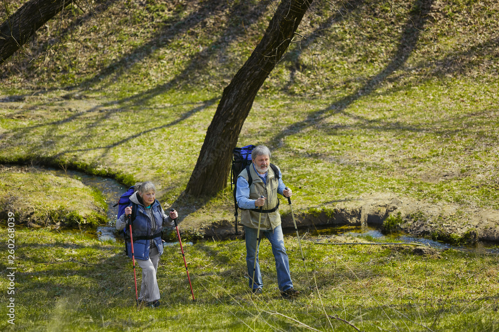 Naklejka premium In way for health and force. Aged family couple of man and woman in tourist outfit walking at green lawn in sunny day near by creek. Concept of tourism, healthy lifestyle, relaxation and togetherness.