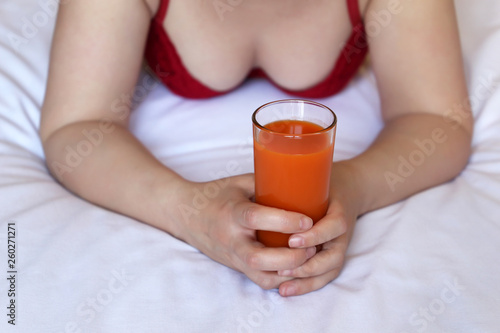 Woman drinks carrot juice, lying on the bed in the morning. Glass of fresh vegetable juice in female hands, concept of healthy diet, body care