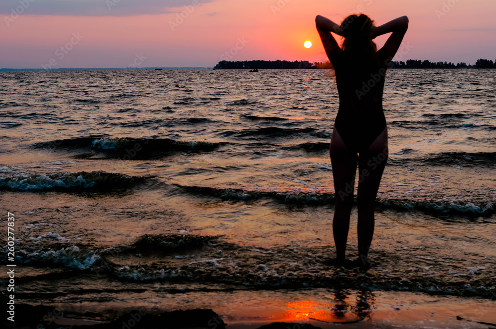 back view of slim woman silhouette in swimsuit with raised arms looking at amazing sunrise near sea on beach