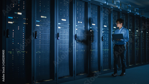 Foto In Dark Data Center: Male IT Specialist Walks along the Row of Operational Server Racks, Uses Laptop for Maintenance