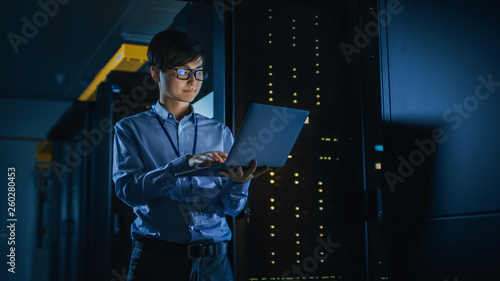 In Dark Data Center: Male IT Specialist Stands Beside the Row of Operational Server Racks, Uses Laptop for Maintenance. Concept for Cloud Computing, Artificial Intelligence, Cybersecurity. Neon Lights photo