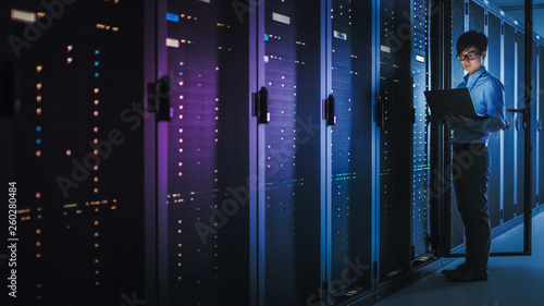 In Dark Data Center: Male IT Specialist Stands Beside the Row of Operational Server Racks, Uses Laptop for Maintenance. Concept for  Artificial Intelligence, Supercomputer, Cybersecurity. Neon Lights photo