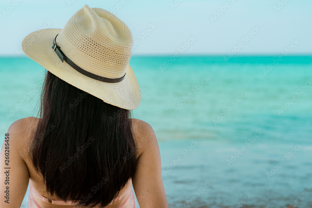Back view of happy young Asian woman in pink swimsuit and straw hat relax and enjoy holiday at tropical paradise beach at sunset. Girl in summer vacation fashion. Beauty sexy model. Summer vibes.