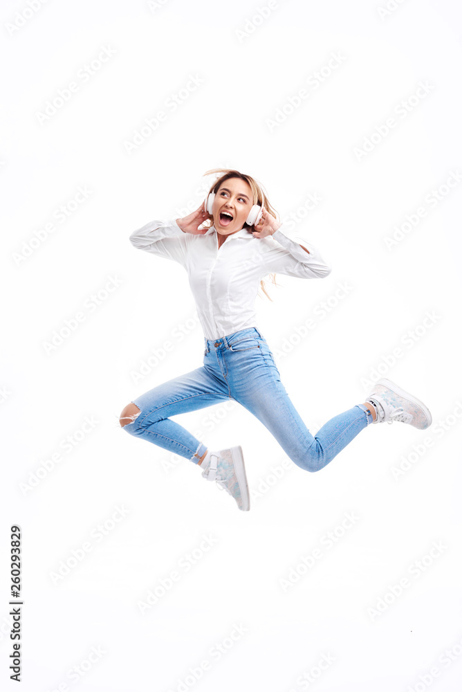 Gorgeous redhead lady listening music on radio in headphones and jumping high. Charming girl in in casual clothes and earphones dancing with hair waving on white background. Full-length portrait