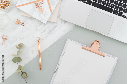 Office desk table with laptop, clipboard, rose gold pen, clips and supplies, pretty eucalyptus branches on marble gray background. Top view with copy space