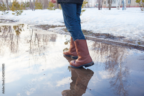 A man in leather boots is on puddles in early spring. Puddles, mud and slush in Russia. Weather and seasons.