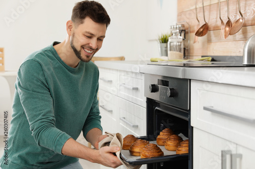 Handsome man taking out tray of baked buns from oven in kitchen