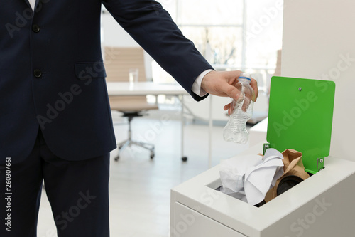Man putting used plastic bottle into trash bin in office, closeup. Waste recycling