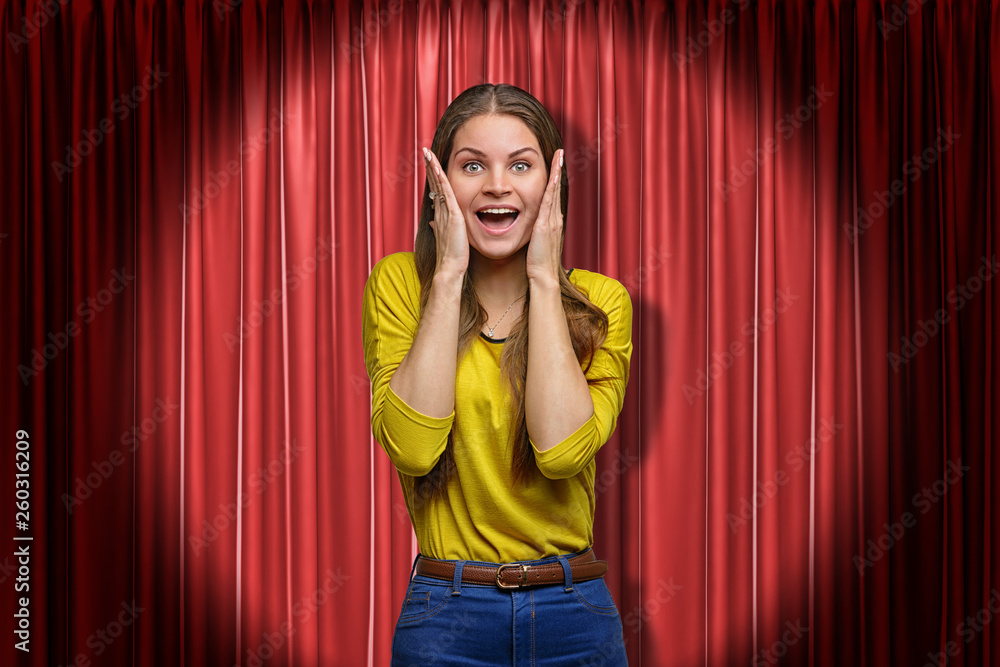 Happy young woman standing and looking at camera with hands on face, in  spotlight, against red stage curtain. Stock Photo | Adobe Stock