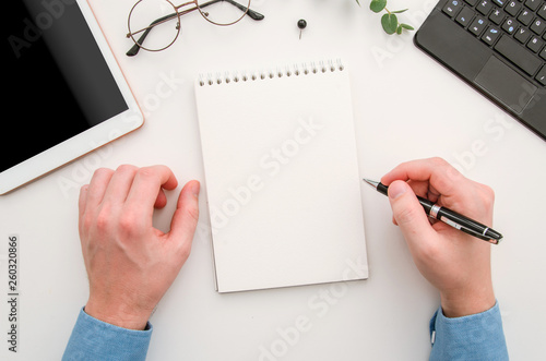 Top view man's hands with notepad on office desk. Workplace with notebook, tablet, glasses, pen and pin. Flat lay man taking notes at workplace