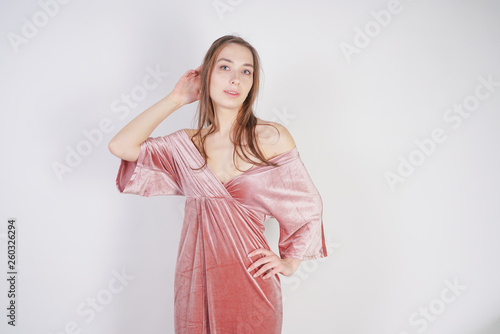 a charming young girl with big eyes and brown hair stands without makeup in a pink pale velvet dress on a white background in the Studio