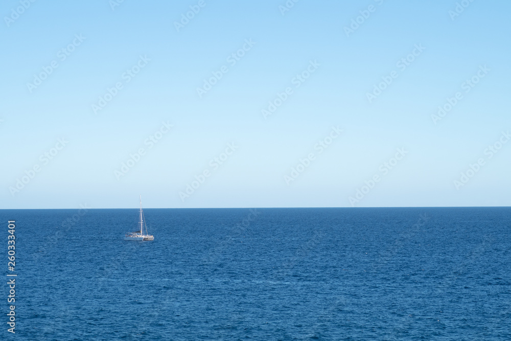 View over a calm Atlantic Ocean with glistening water, palm leafs and one single sailboat passing by.