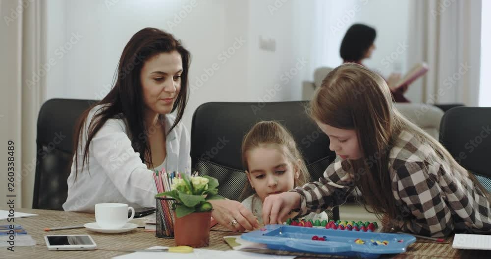 Smiling large mature mother with her two daughters playing together on table games spending a family time together , grandmother reading a book on the background sitting on the sofa. 4k Smiling large mature mother with her two daughters playing together on table games spending a family time together , grandmother reading a book on the background sitting on the sofa. 4k