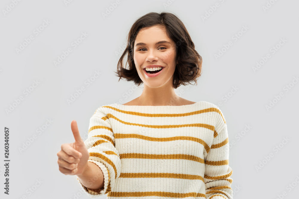 gesture and people concept - happy smiling young woman in striped pullover showing thumbs up over grey background gesture and people concept - happy smiling young woman in striped pullover showing thumbs up over grey background