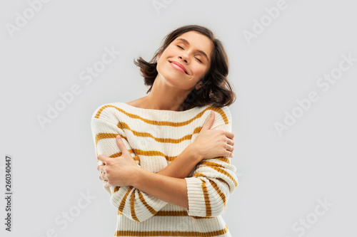 people concept - happy pleased young woman in striped pullover with closed eyes hugging herself over grey background