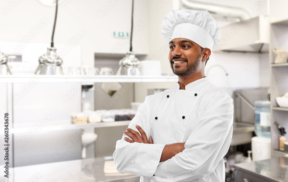 cooking, profession and people concept - happy male indian chef in toque with crossed arms over restaurant kitchen background