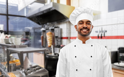 cooking, profession and people concept - happy male indian chef in toque over kebab shop kitchen background